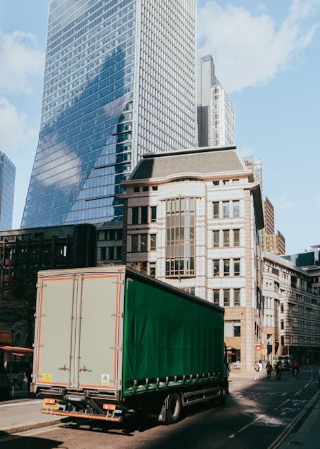 A large commercial box truck with a green cargo cover is parked on a city street, in front of modern glass office buildings and older stone and brick structures. The truck is positioned near the pavem