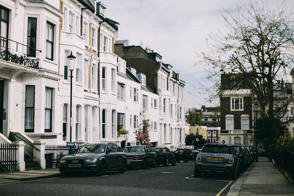 A residential street with white multi-storey terraced houses featuring bay windows and decorative cornices. Several cars, including a grey Audi and a Mini, are parked along the pavement, occupying des