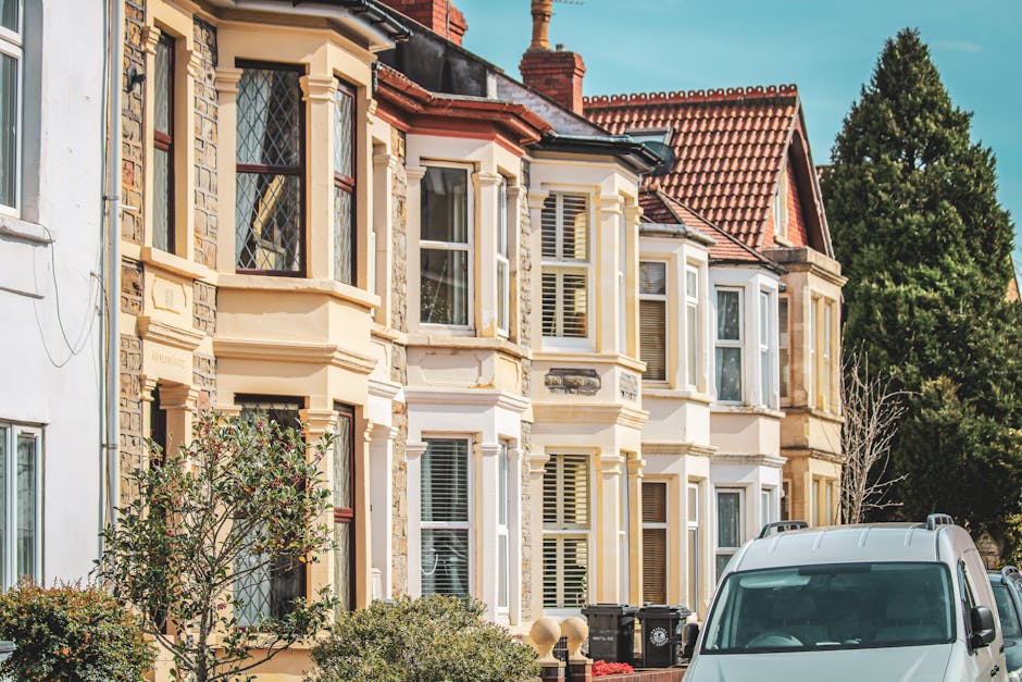 A row of Victorian terraced houses constructed with grey stone and brick, featuring gabled roofs, chimney stacks, and white-framed sash windows, situated behind a well-maintained green lawn and leafle